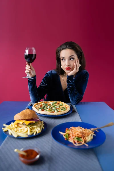 Bruna donna con bicchiere di vino guardando lontano vicino a pizza e piatti con gustoso pasto servito sul tavolo blu isolato sul rosso — Foto stock