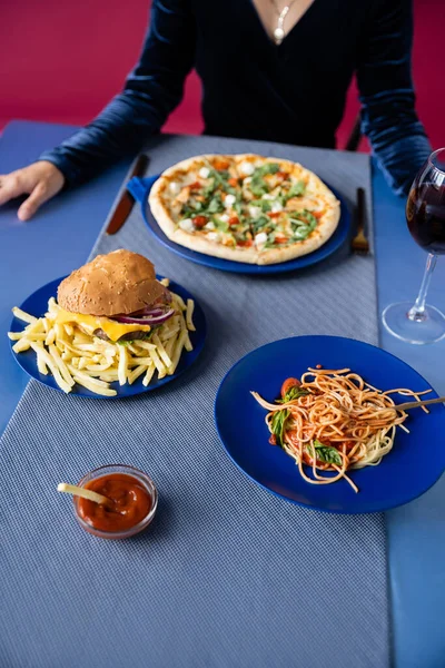 Vista ritagliata di donna sfocata vicino hamburger con patatine fritte, pizza e pasta isolata su rosso — Foto stock