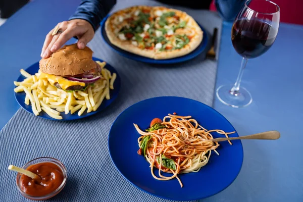 Partial view of woman taking burger near wine glass and plates with pasta and pizza on blue table — Stock Photo