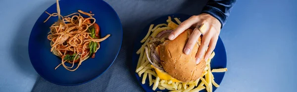 Vista superior de la mujer recortada tomando hamburguesa cerca de papas fritas y espaguetis en platos azules, pancarta - foto de stock