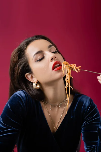 Brunette woman in blue dress enjoying tasty pasta with closed eyes isolated on red — Stock Photo