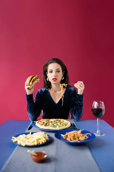 Mujer elegante con hamburguesa y espaguetis cerca de la mesa azul con papas fritas y copa de vino aislado en rojo - foto de stock