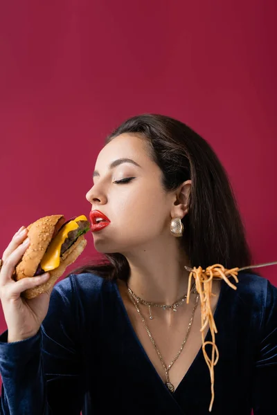 Elegante morena mujer con sabrosa hamburguesa y espaguetis en tenedor aislado en rojo - foto de stock