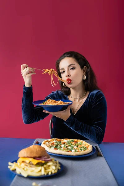 Стильная женщина в велюровом платье ест спагетти рядом с бургером с картошкой фри и пиццей, изолированной на красном — стоковое фото