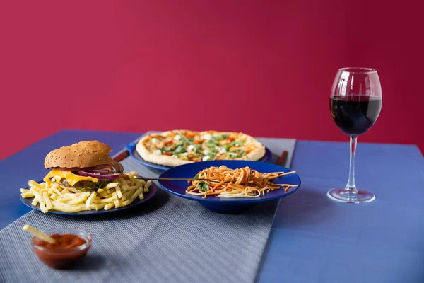 Pratos azuis com massa e pizza borrada perto de batatas fritas com hambúrguer e copo de vinho isolado em vermelho — Fotografia de Stock