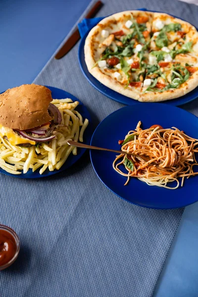 Vista superior de espaguetis cerca de papas fritas con hamburguesa y pizza borrosa en platos azules - foto de stock