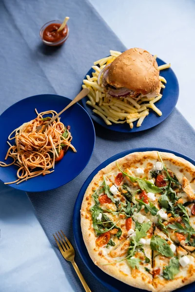 Top view of french fries with burger near tasty pasta and pizza on blue plates — Stock Photo