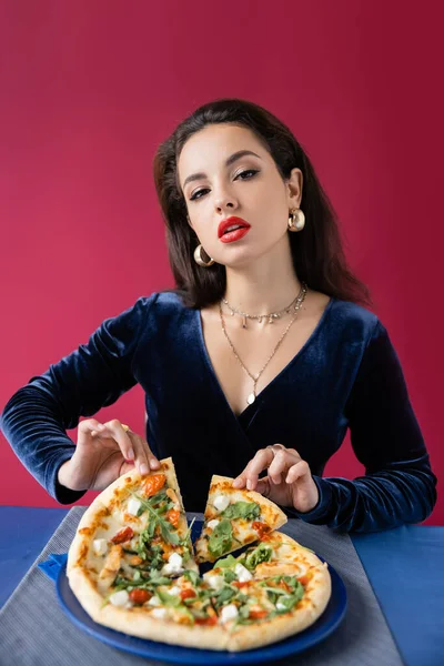 Чувственная женщина в синем велюровом платье и золотых аксессуаров принимая кусок вкусной пиццы изолированы на красный — стоковое фото