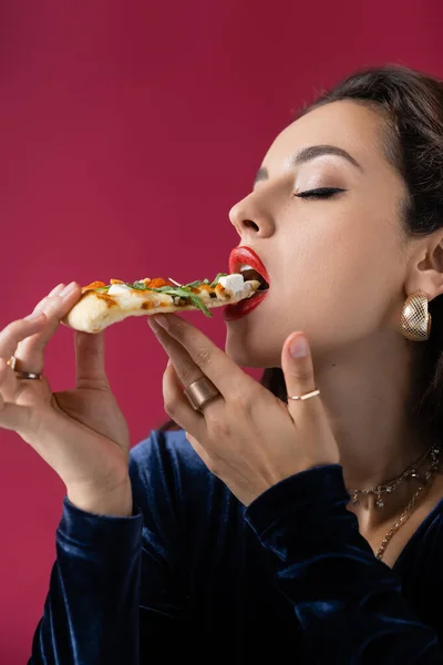 Mujer elegante y sexy en anillos de dedo de oro comiendo deliciosa pizza aislada en rojo - foto de stock