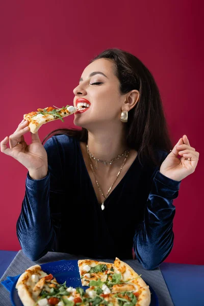 Mujer sensual en vestido de terciopelo azul comiendo sabrosa pizza aislada en rojo - foto de stock