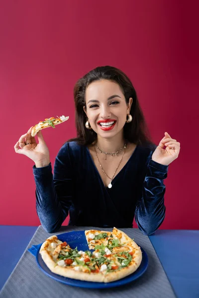 Freudige Frau im Samtkleid hält Pizza in der Hand und blickt in die Kamera neben blauem Tisch isoliert auf rotem — Stockfoto