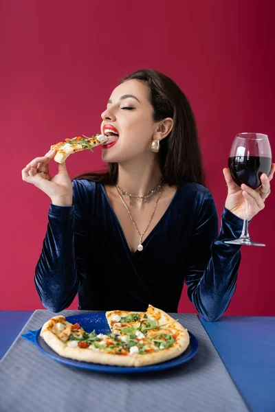 Mujer bonita y elegante con copa de vino comiendo sabrosa pizza aislada en rojo - foto de stock