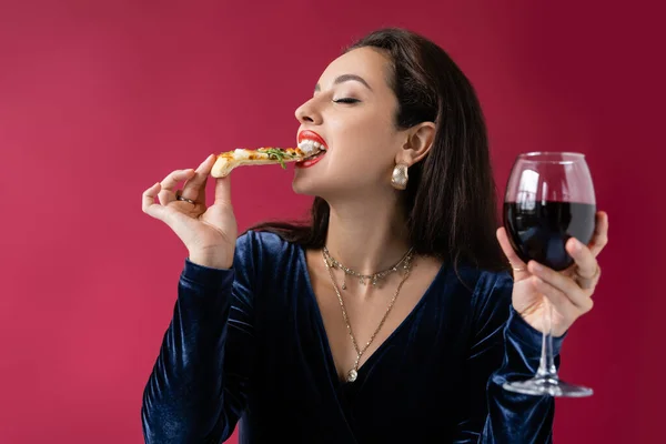 Elegante donna bruna con bicchiere di vino mangiare pizza italiana isolata sul rosso — Foto stock