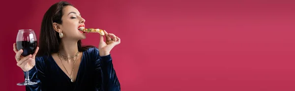 Mujer joven en vestido de terciopelo azul sosteniendo vaso de vino y comiendo sabrosa pizza aislada en rojo, bandera - foto de stock