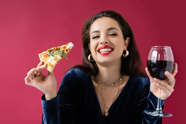 Femme heureuse en robe de velours bleu et boucles d'oreilles dorées tenant vin et pizza isolé sur rouge — Photo de stock