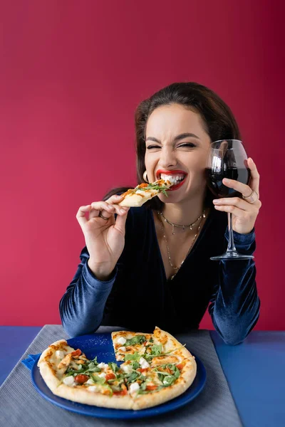 Fröhliche Frau mit einem Glas Wein, die leckere Pizza in der Nähe des blauen Tisches isst, isoliert auf rot — Stockfoto