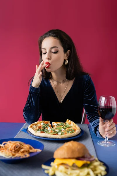 Femme sexy avec un verre de vin léchant son doigt près de la table bleue servi avec un délicieux repas isolé sur rouge — Photo de stock