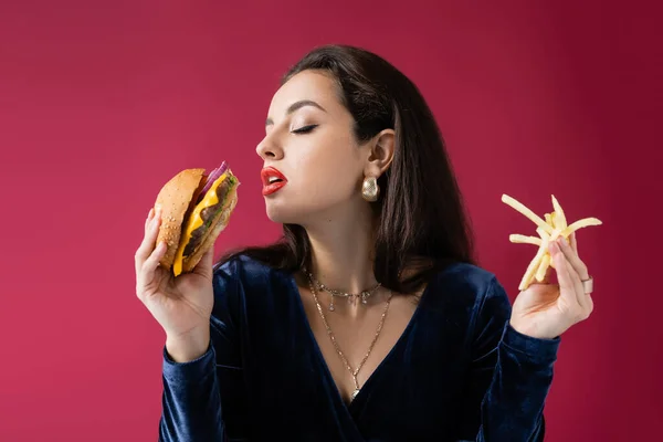 Young woman in blue velour dress holding burger and french fries isolated on red — Stock Photo