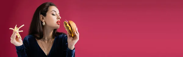 Mulher sensual em vestido elegante segurando batatas fritas e delicioso hambúrguer isolado em vermelho, banner — Fotografia de Stock