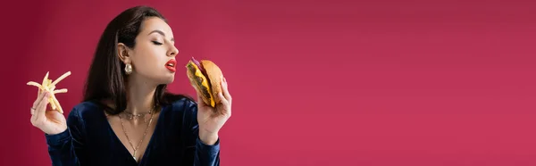 Mujer sensual en vestido elegante sosteniendo papas fritas y deliciosa hamburguesa aislada en rojo, bandera - foto de stock