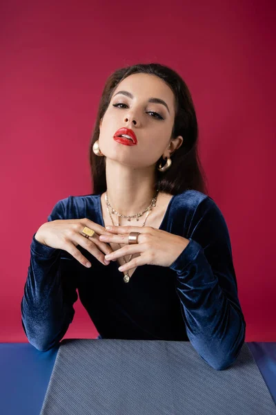 Mujer glamour en elegante vestido de terciopelo mirando a la cámara cerca de la mesa con mantel azul aislado en rojo - foto de stock