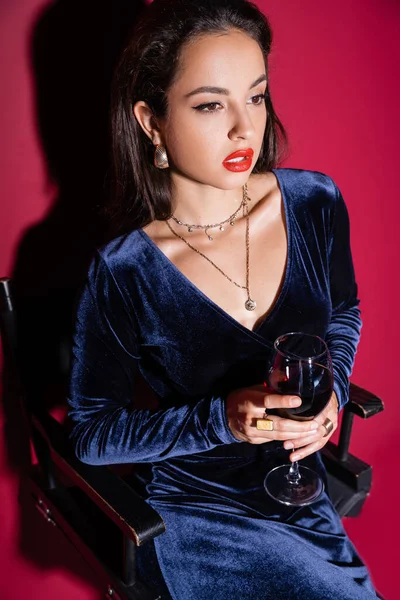 Чувственная женщина в синем велюровом платье сидит с бокалом вина на красном фоне — стоковое фото