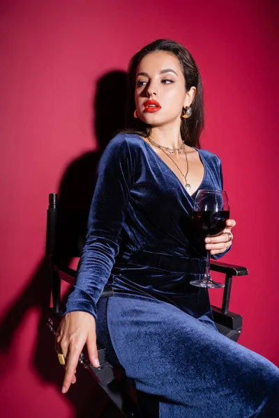 Femme séduisante en robe élégante assise avec un verre de vin et regardant loin sur fond rouge — Photo de stock