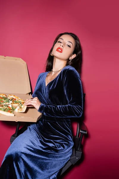 Donna seducente in abito di velluto blu che tiene scatola di cartone con pizza mentre guarda la fotocamera su sfondo rosso — Foto stock