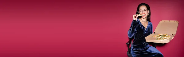 Pleased woman sitting with carton box of pizza and eating on red background, banner — Stock Photo