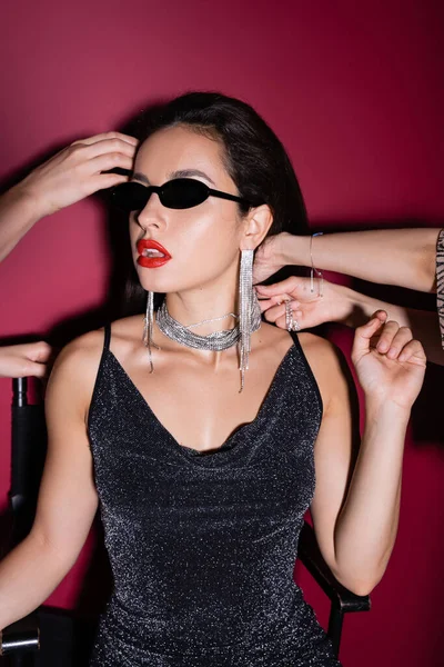 Sexy woman in sunglasses and with red lips near hands touching her on burgundy background — Stock Photo