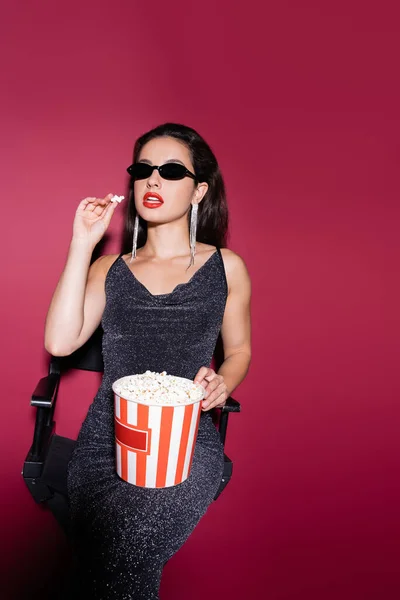 Young woman in black sunglasses and lurex dress eating popcorn on red background — Stock Photo