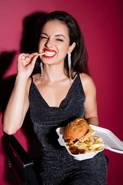 Donna allegra in abito elegante nero mangiare patatine fritte vicino hamburger in contenitore di plastica su sfondo rosso — Foto stock