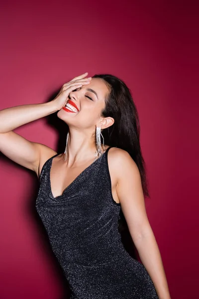 Elegant woman in shiny earrings and black lurex dress obscuring face and laughing on red background — Stock Photo