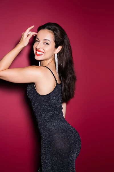 Glückliche brünette Frau im schwarzen Lurexkleid lächelt in die Kamera auf rotem Hintergrund — Stockfoto
