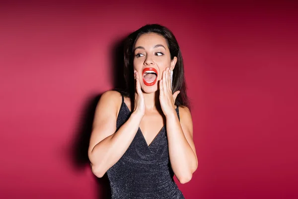 Scared woman in elegant dress touching face and screaming on red background — Stock Photo
