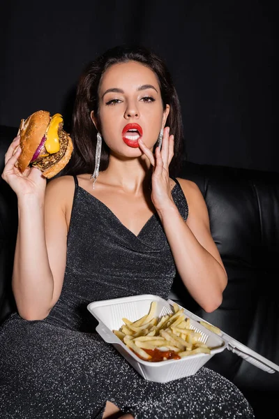 Donna seducente con gustoso hamburger e patatine fritte toccare labbro e guardando la fotocamera isolata su nero — Foto stock