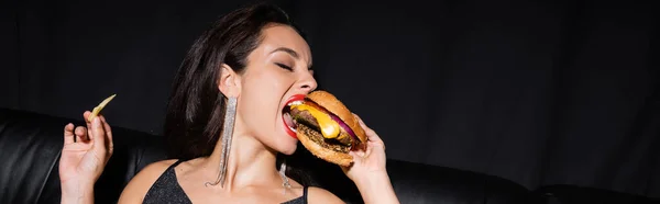 Morena mujer con los ojos cerrados comiendo sabrosa hamburguesa aislado en negro, pancarta - foto de stock