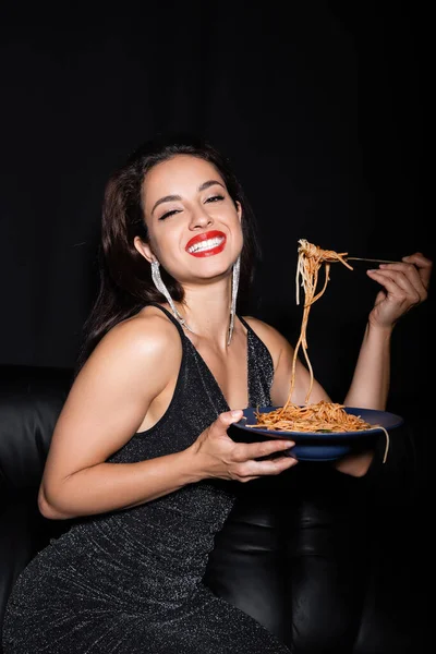 Femme gaie et élégante avec assiette de pâtes savoureuses souriant à la caméra isolée sur noir — Photo de stock