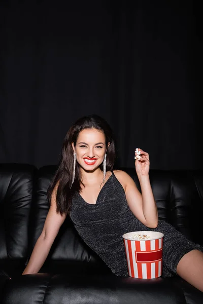 Donna allegra guardando macchina fotografica vicino secchio di carta di popcorn isolato su nero — Foto stock