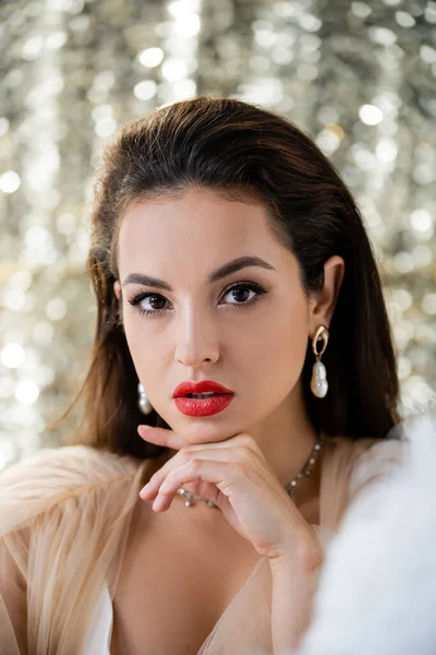 Morena mujer con maquillaje y pendientes de perlas sosteniendo la mano cerca de la barbilla y mirando a la cámara en un fondo brillante - foto de stock