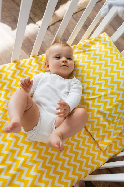 Top view of barefoot infant girl in baby romper lying on yellow bedding in crib — Stock Photo