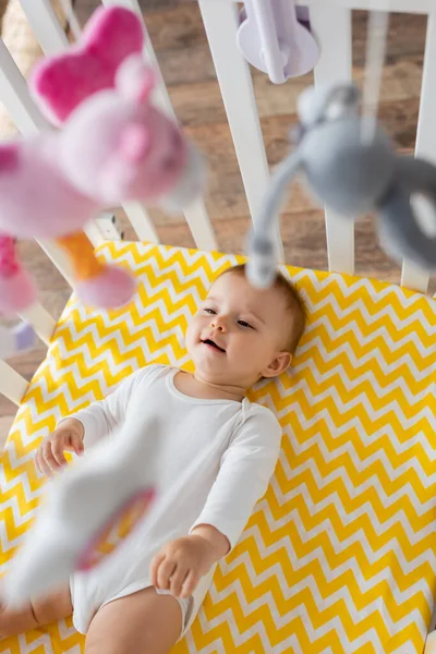 Top view of happy infant girl in baby romper lying in crib and looking at hanging mobile with soft toys — Stock Photo