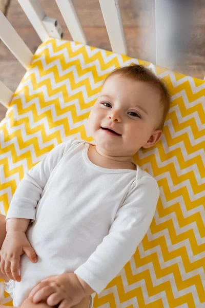 Top view of happy infant girl in baby romper lying on yellow bedding in crib — Stock Photo
