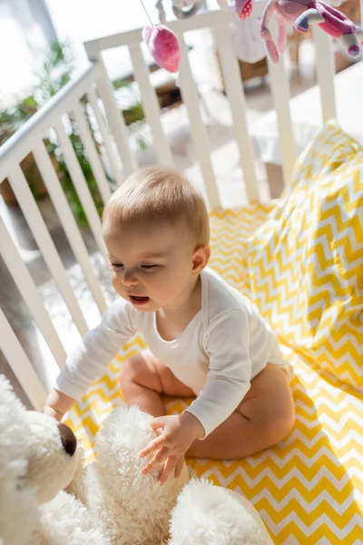 High angle view of cute infant girl in baby romper sitting near teddy bear in crib — Stock Photo