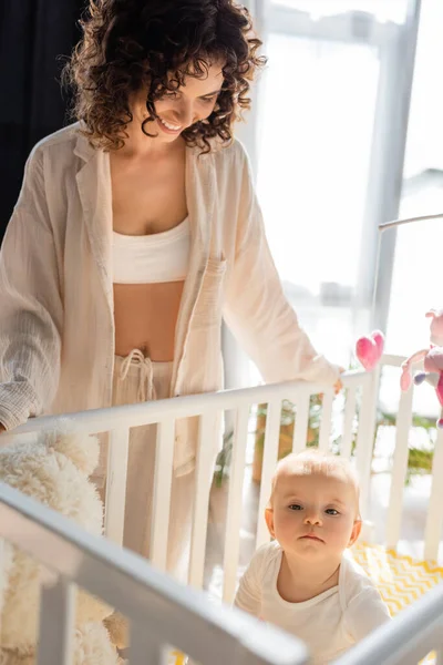 Mujer rizada en ropa de salón sonriendo y mirando a la hija bebé en cuna - foto de stock