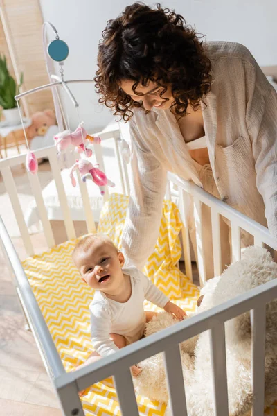 Visão de alto ângulo de mãe alegre em loungewear olhando para a filha do bebê sentado perto de ursinho de pelúcia no berço — Fotografia de Stock