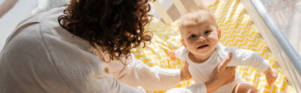 High angle view of curly mother in loungewear taking baby daughter in romper from crib, banner — Stock Photo