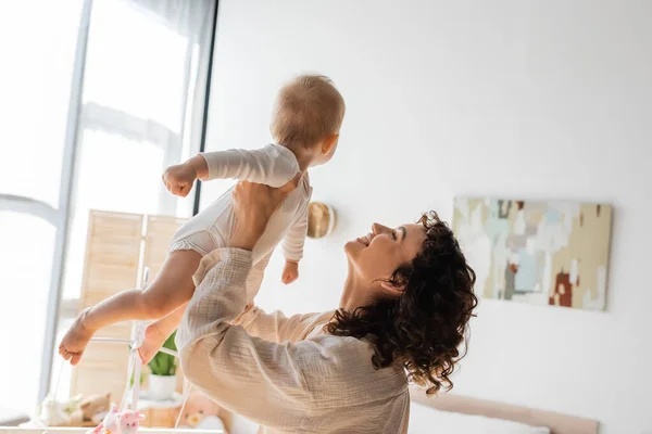 Feliz y rizada madre en ropa de salón sosteniendo en brazos bebé hija en mamada - foto de stock