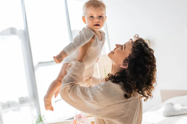 Happy and curly woman in loungewear holding in arms barefoot daughter in romper — Stock Photo