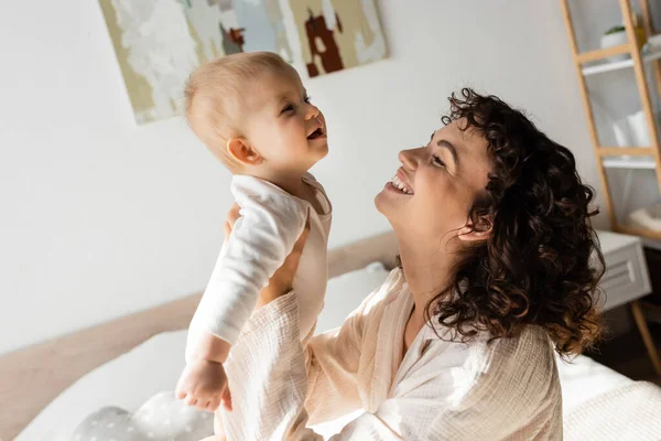 Mujer alegre y rizada en ropa de salón sosteniendo en brazos bebé hija en mameluco - foto de stock